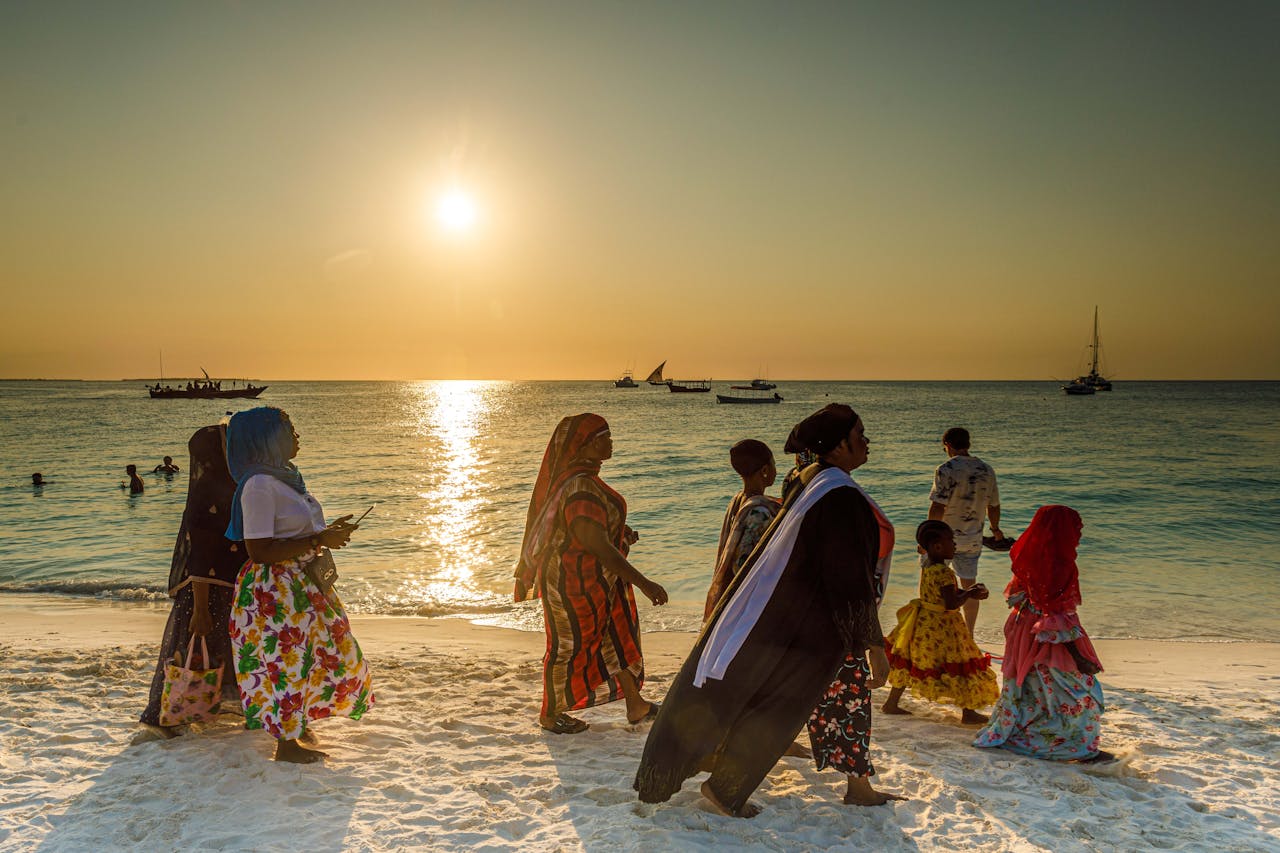 A group of people in colorful attire walk along a beach with a vibrant sunset.