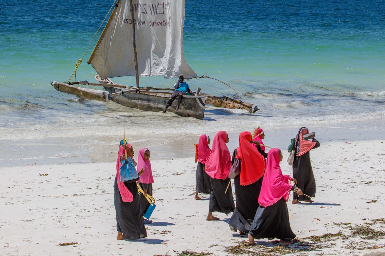 Women in colorful attire walking on a beach with a traditional boat in the background.