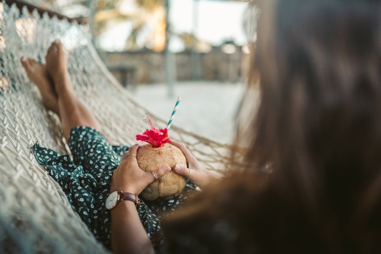 A woman enjoys a coconut drink while relaxing in a hammock on a sandy beach.